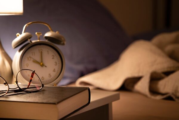 Alarm clock, reading glasses, and a book on a nightstand beside a bed at night β better sleep supports fat loss