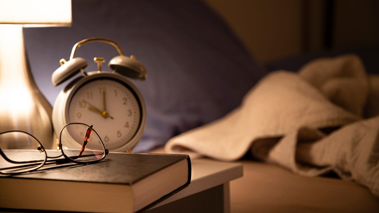 Alarm clock, reading glasses, and a book on a nightstand beside a bed at night β better sleep supports fat loss